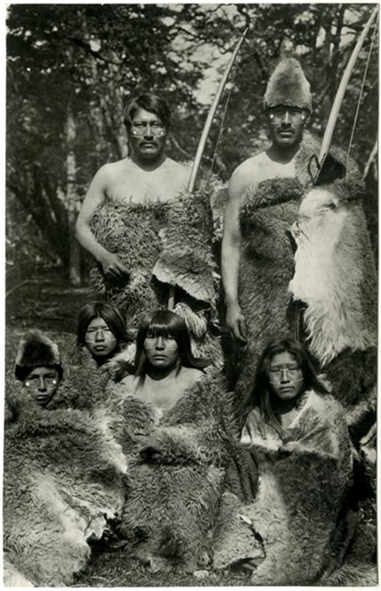 Grupo de mujeres y hombres selk’nam vistiendo pieles de guanaco y portando arcos de caza.